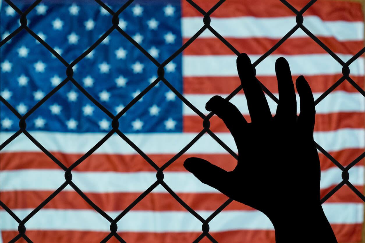 A silhouette of a hand reaching onto a chainlink fence while a well-lit American flag is in the background.